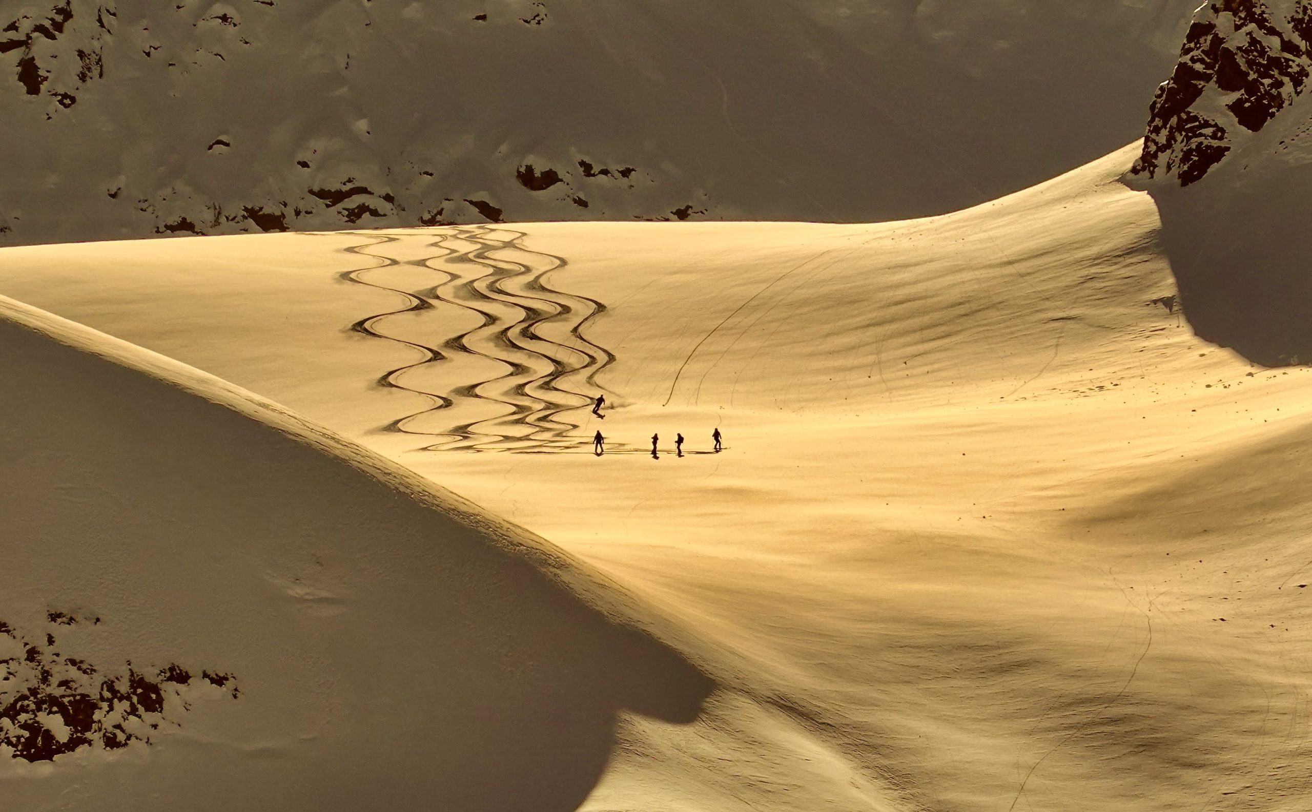 Ski tracks wind down a golden, sunlit snowfield with skiers below